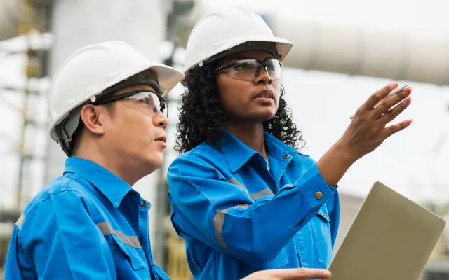 Two engineers in blue safety uniforms and white helmets discussing while one gestures and holds a laptop outdoors.