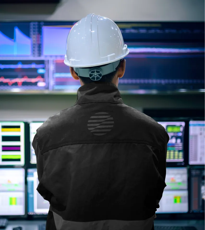 Engineer wearing a white hard hat and dark jacket monitors multiple data screens in a control room.