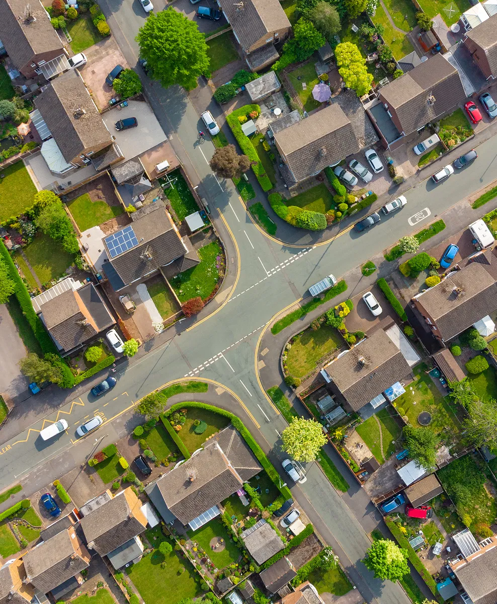 Aerial view of a residential neighborhood with an intersection surrounded by houses, gardens, and parked cars.