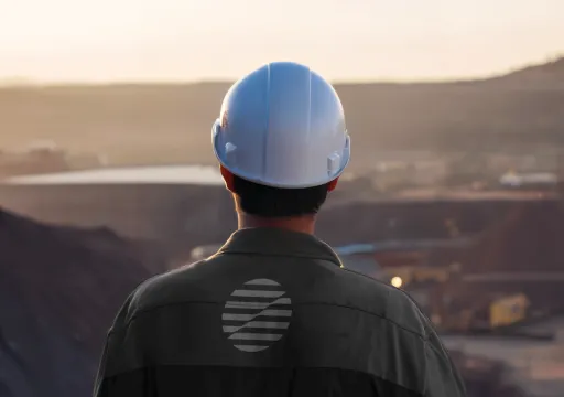 Worker wearing a white hard hat overlooking an industrial mining site at sunset.