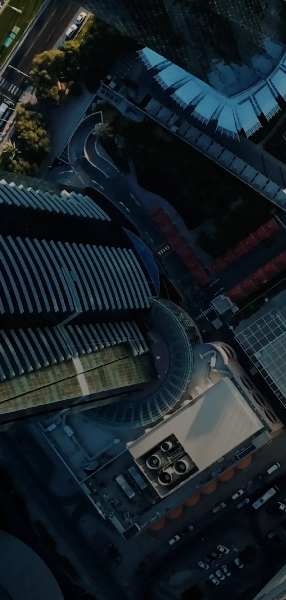 Aerial view of urban area with tall modern buildings, curved roads, parked cars, and trees in soft evening light.