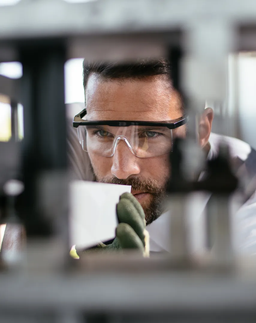 Man wearing safety glasses and gloves closely inspecting a small white object in an industrial setting.