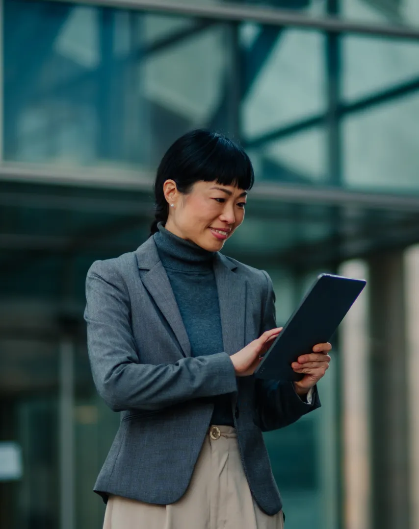 Smiling woman in gray blazer using a tablet outdoors with a modern glass building in the background.