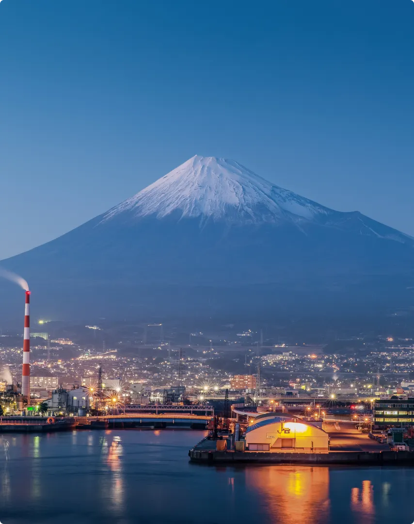 Snow-capped Mount Fuji towering over a cityscape at dusk with illuminated buildings and a waterfront industrial area.