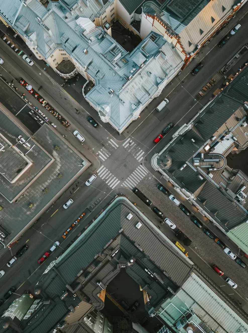 Aerial view of an urban intersection with crosswalks, surrounded by tall buildings with varied rooftops and parked cars along the streets.