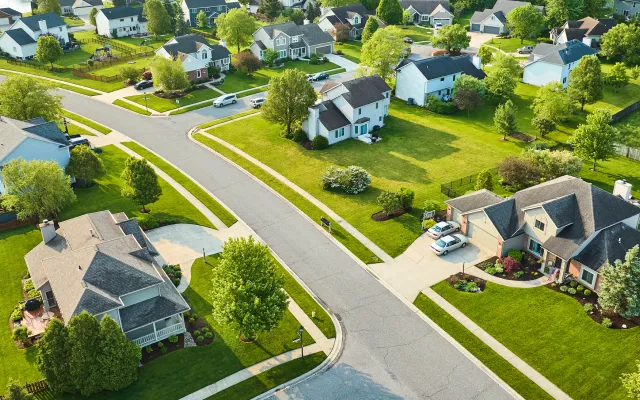 Aerial view of a suburban neighborhood with detached houses, green lawns, trees, and curved streets on a sunny day.