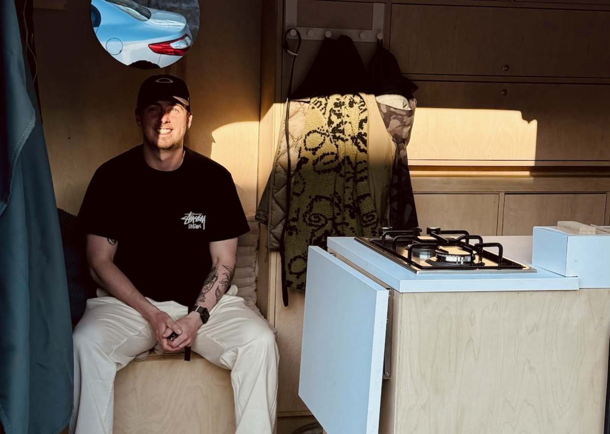 Man wearing a black cap and black t-shirt with white pants sitting inside a wooden camper van kitchen area with coats hanging on the wall behind him.