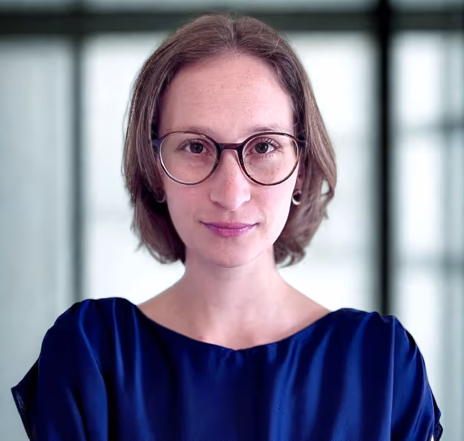 Porträt einer Frau mit kurzen braunen Haaren, großen runden Brillen und blauem Oberteil vor unscharfem Hintergrund.