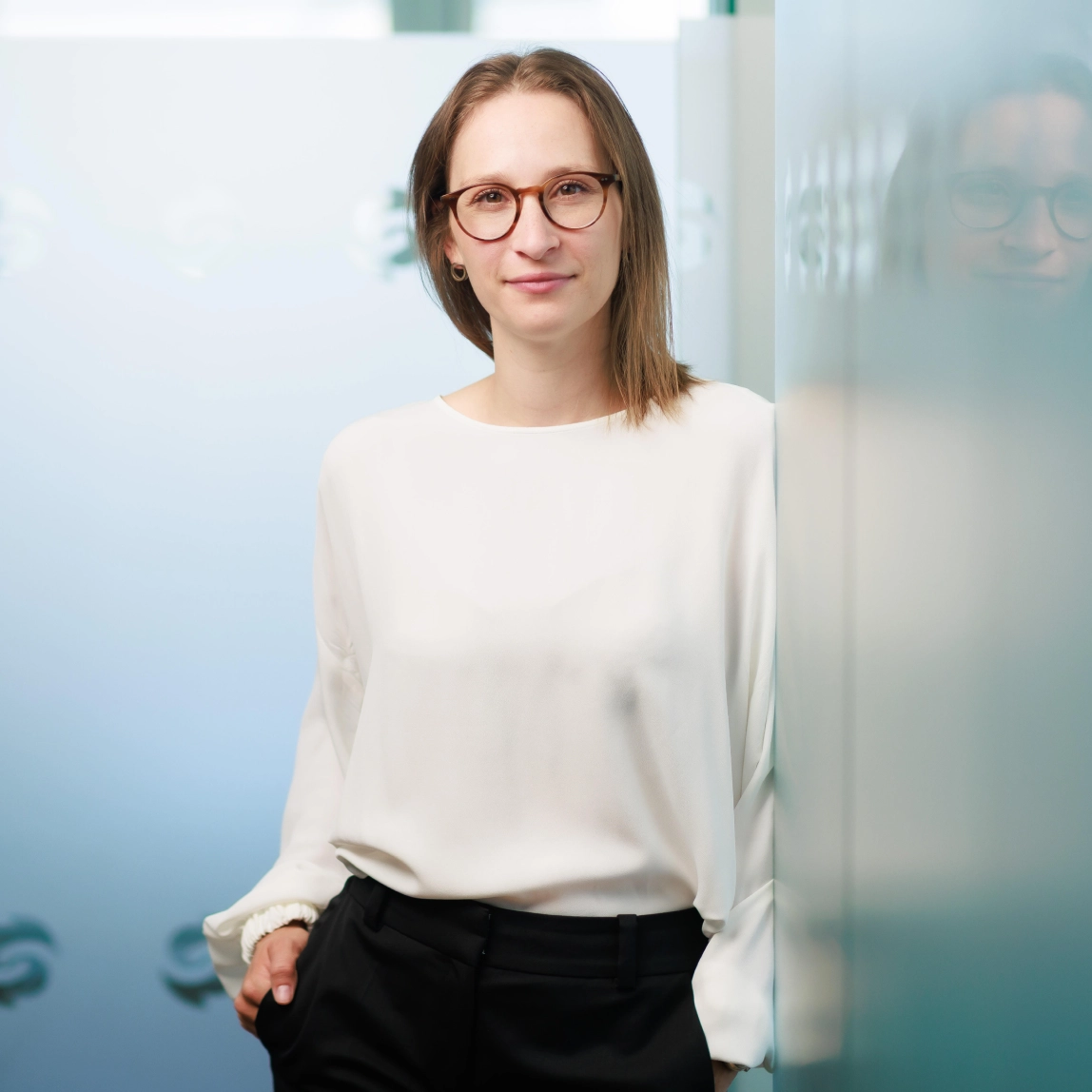 Frau mit kurzen braunen Haaren, trägt eine Brille und ein dunkles Oberteil, vor unscharfem Fensterhintergrund.