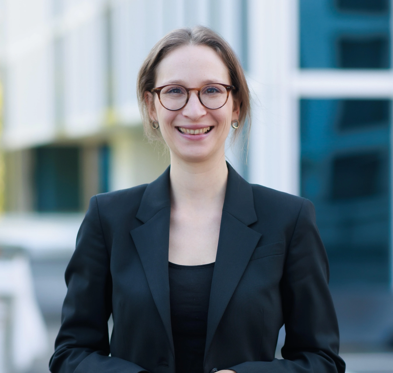 Porträt einer Frau mit kurzen braunen Haaren, großen runden Brillen und blauem Oberteil vor unscharfem Hintergrund.