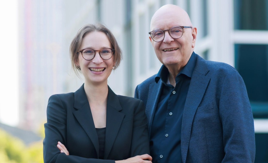 Porträt eines jungen Mannes mit Brille und eines älteren Mannes im Anzug, beide lächeln leicht.
