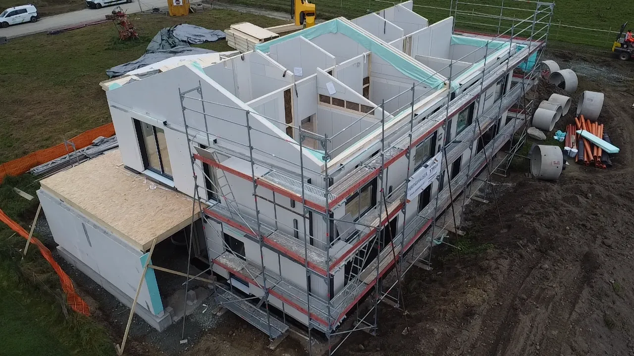 Zweistöckiges Haus im Bau mit Gerüst, weißen Wänden und angrenzender Terrasse auf einem feldnahen Grundstück.