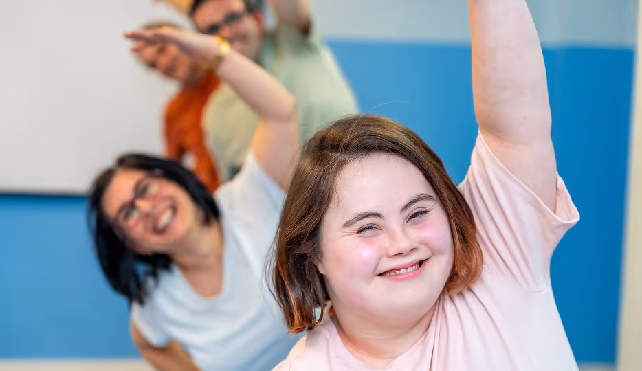 A joyful young woman with and two people behind her smiling and raising one arm in a blue room.