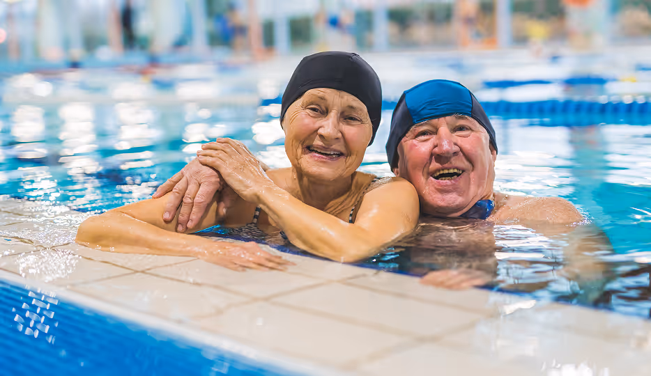 Smiling elderly couple in swim caps relaxing at the edge of an indoor swimming pool.