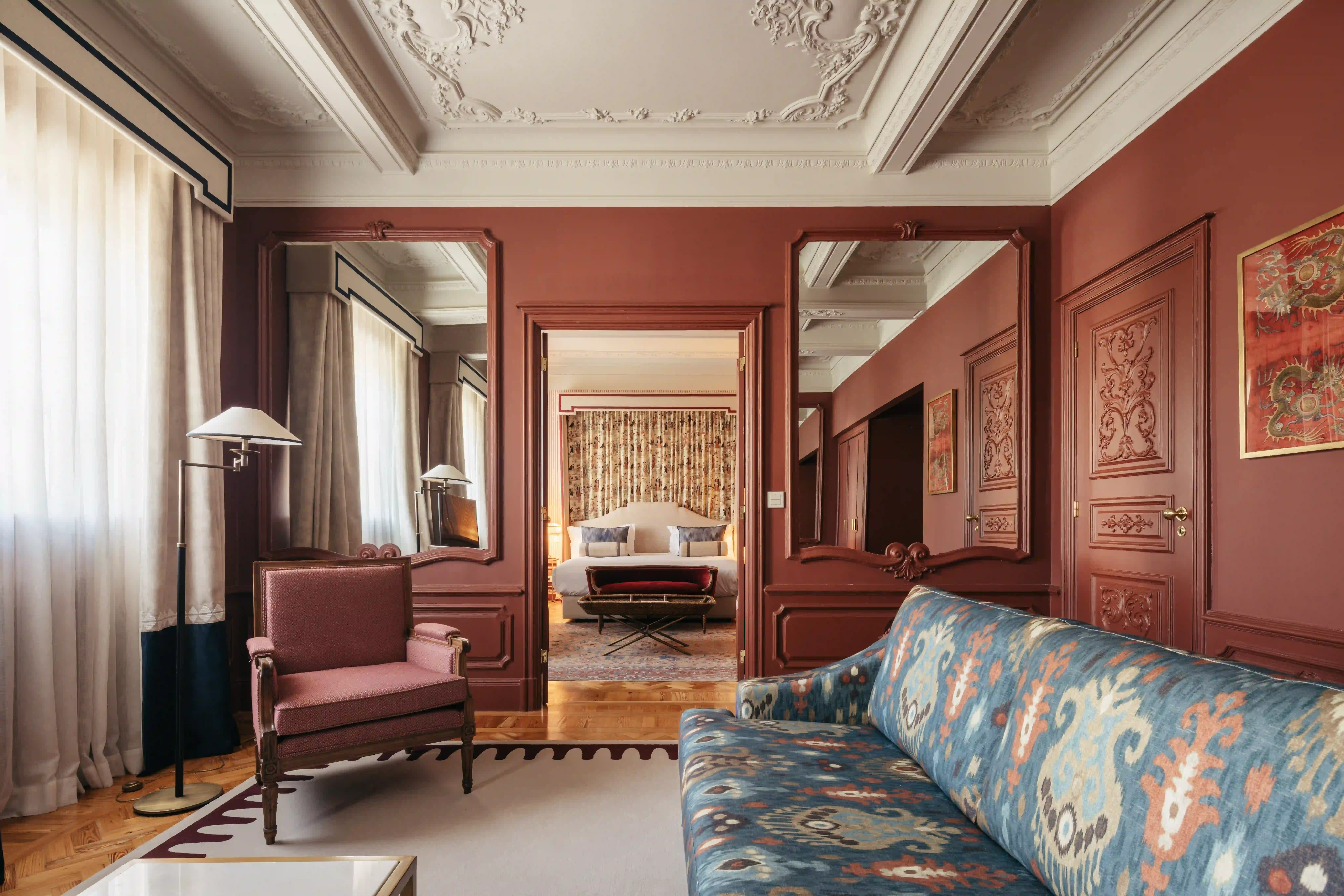 Elegante sala de estar con sofá azul estampado, sillón rojo, techo adornado y puerta que conduce a un dormitorio con cama blanca y cortinas florales.