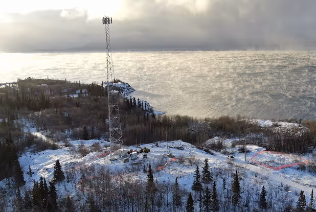 Snow-covered landscape with evergreen trees and a tall communication tower near a large body of water under a cloudy sky.