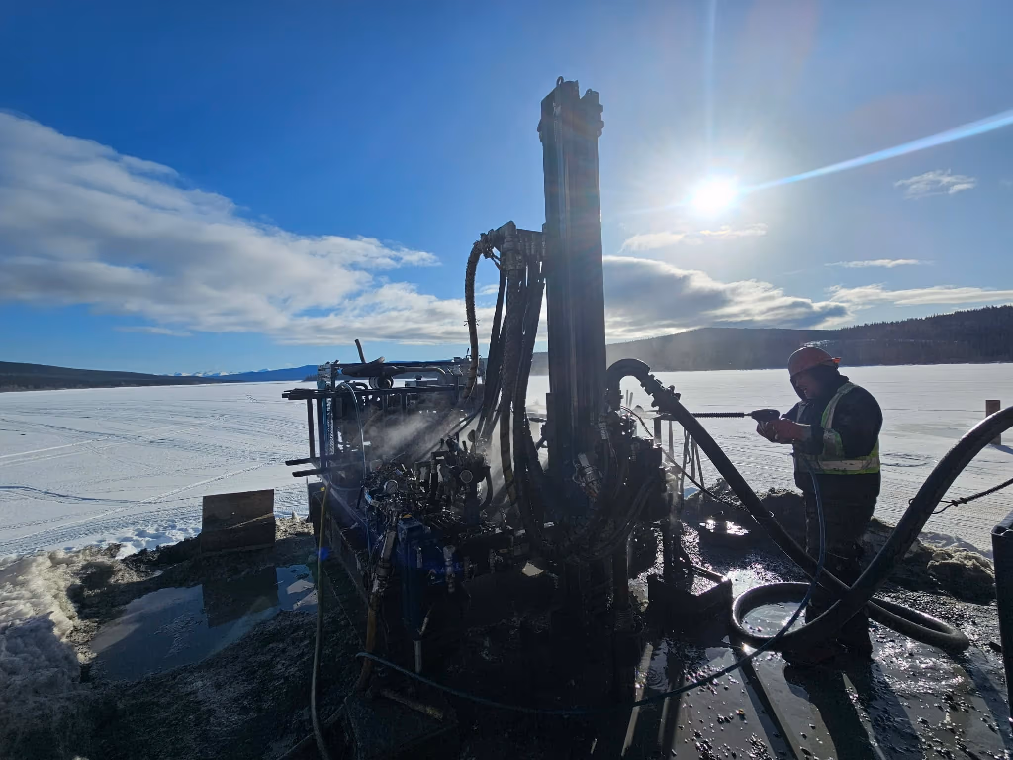Worker in high visibility vest and helmet operating drilling machinery on a snowy landscape under bright sun.
