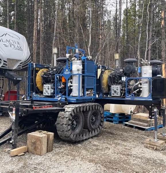 Heavy blue industrial equipment with large tracked wheels mounted on a trailer, set outdoors near a forest.