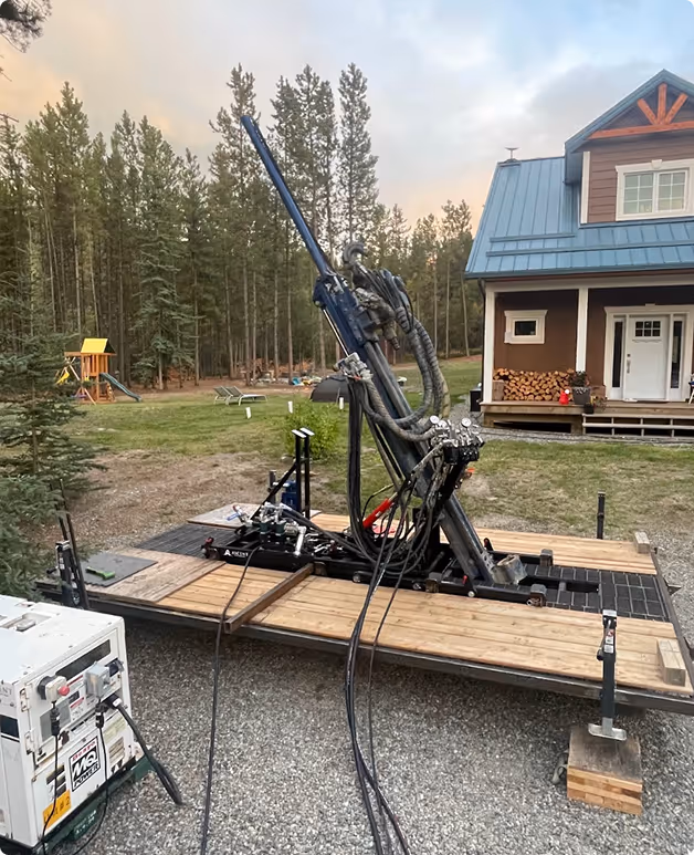 A mounted mechanical device with hoses and cables on a wooden platform in a backyard with a house, forest, and children's playground in the background.