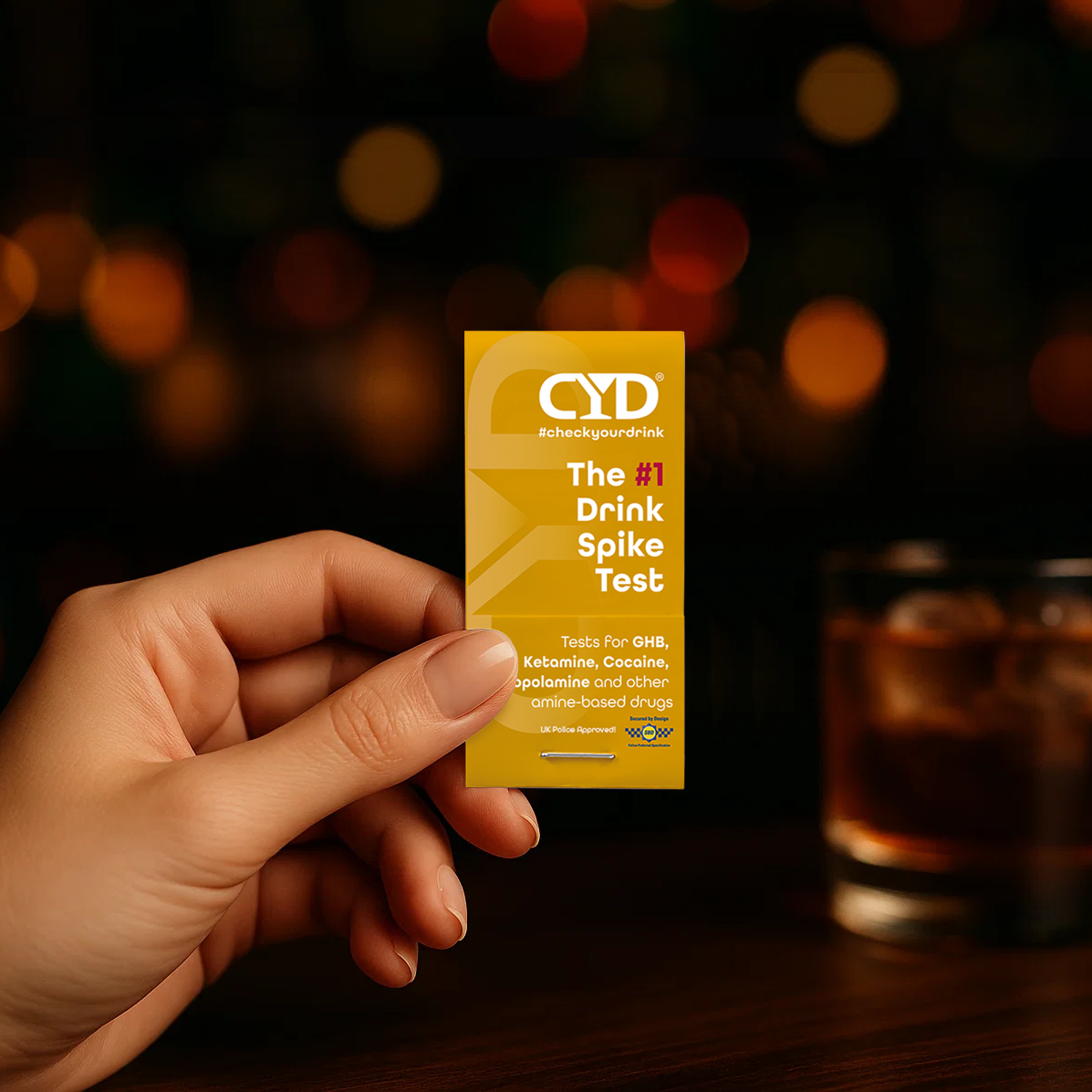 Hand holding a yellow CYD drink spike test packet with a blurred drink in the background.