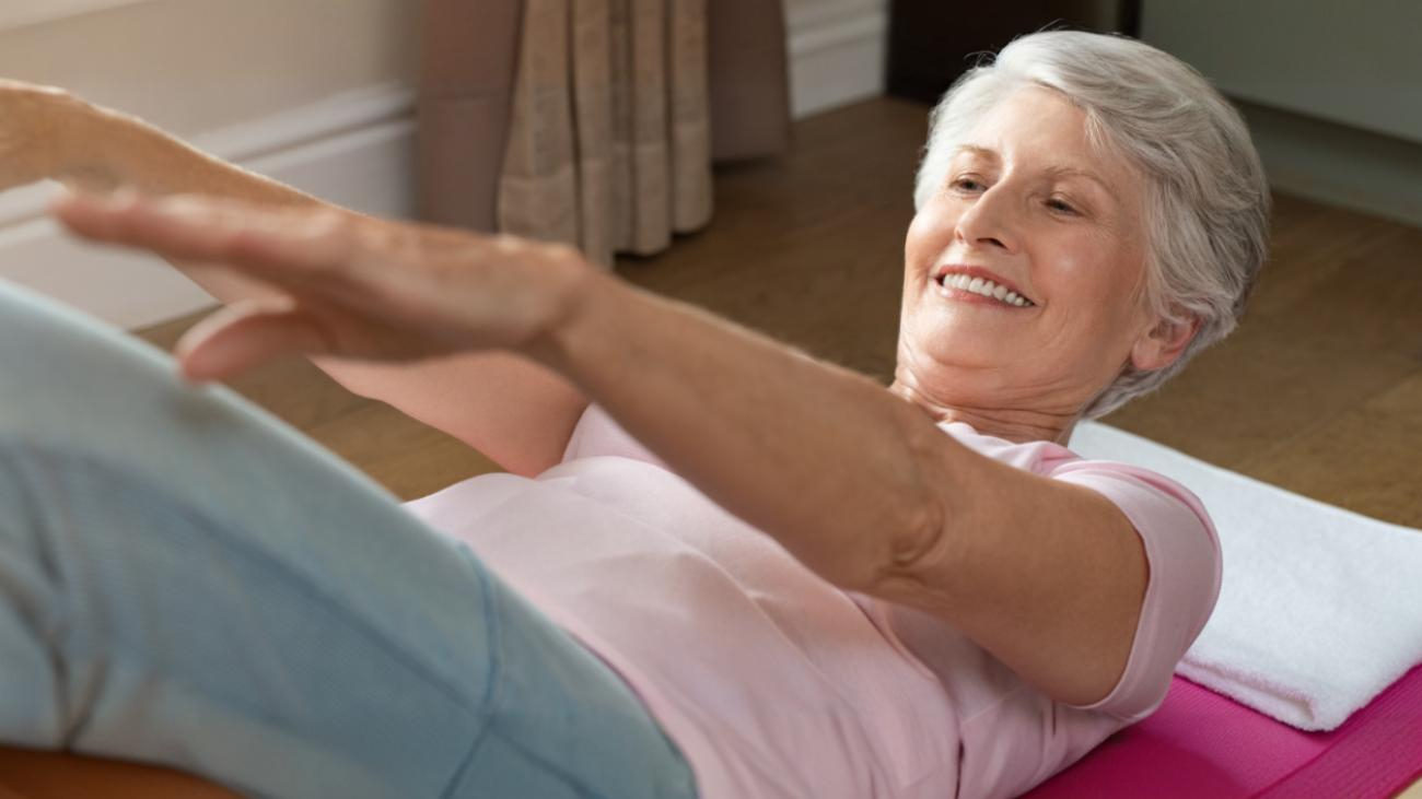 Hymyilevä ikääntynyt nainen harjoittelee fysioterapiaa matolla.