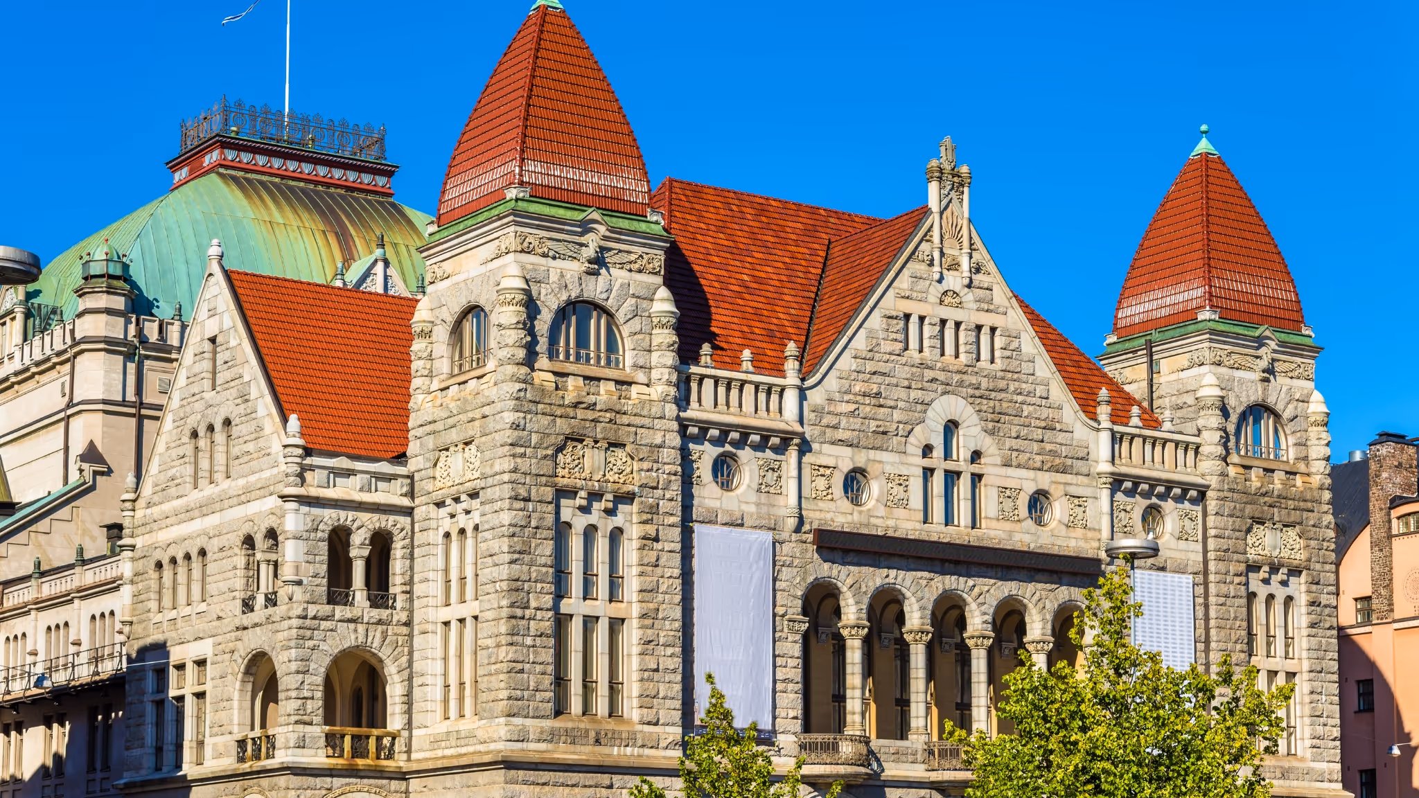 Vanhan harmaasta kivestä rakennetun rakennuksen julkisivu, jossa punaiset tiilikattoiset tornit ja kirkas sininen taivas.