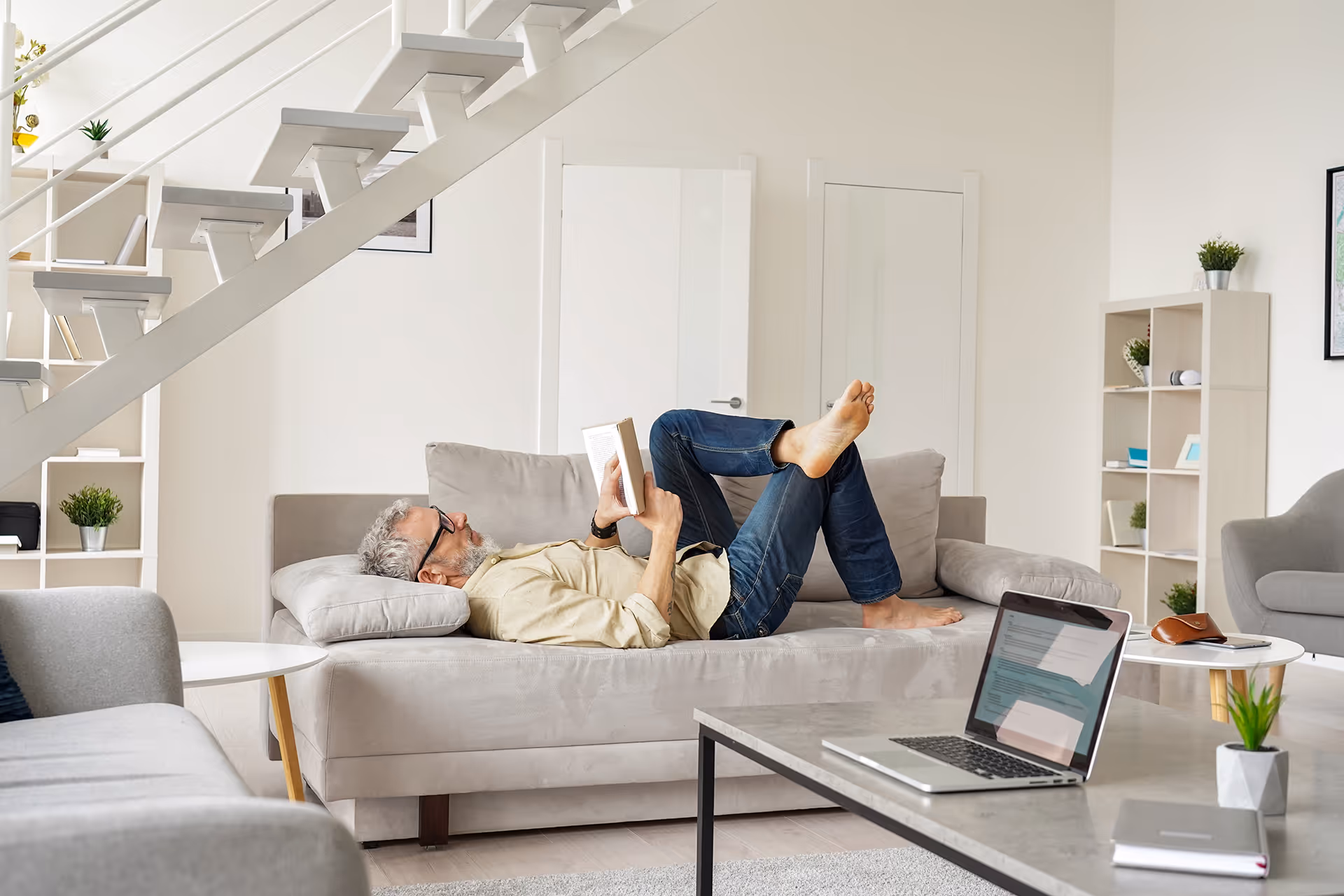 Keski-ikäinen mies rentoutuu sohvalla lukien kirjaa kodikkaassa olohuoneessa.