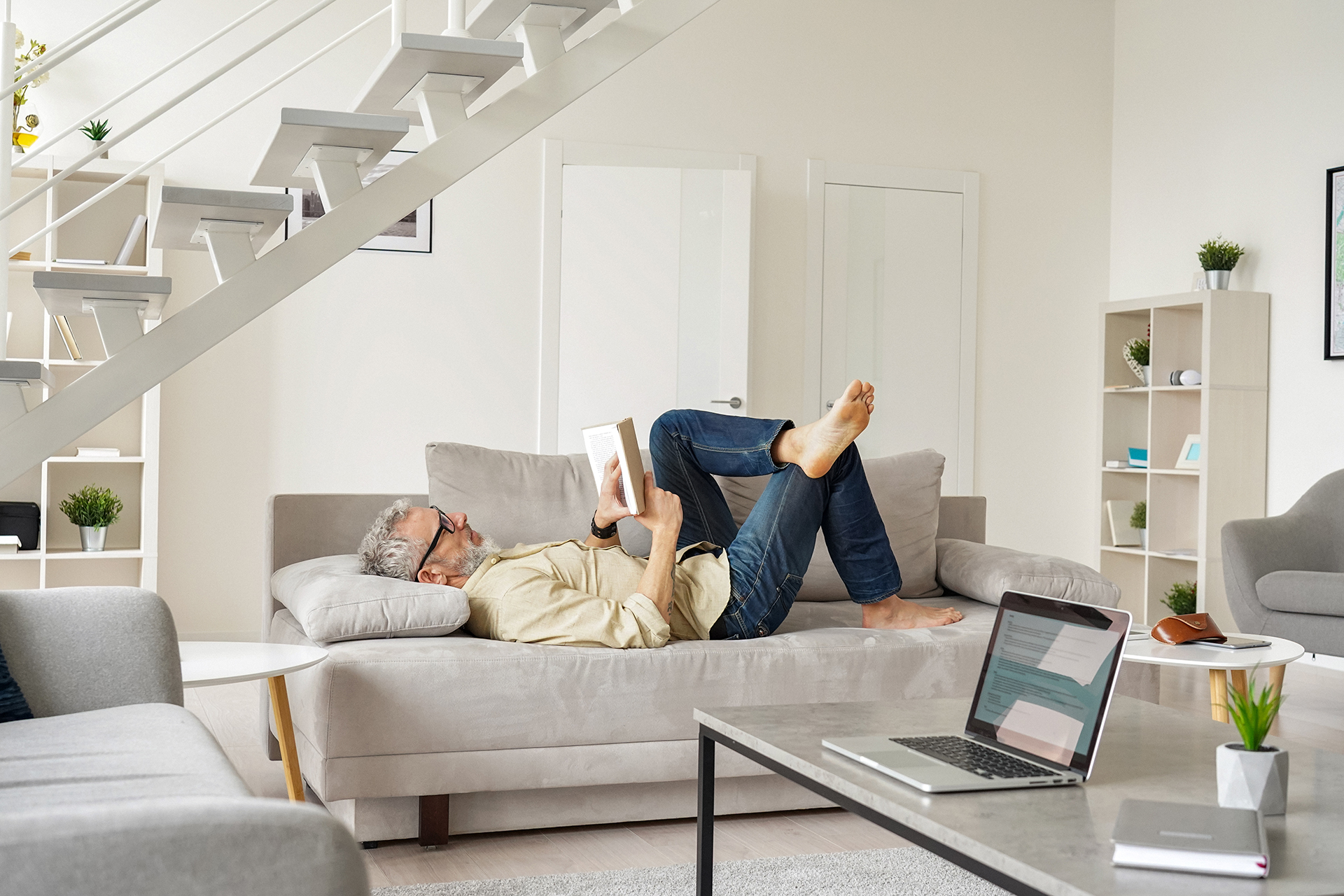 Keski-ikäinen mies rentoutuu sohvalla lukien kirjaa kodikkaassa olohuoneessa.