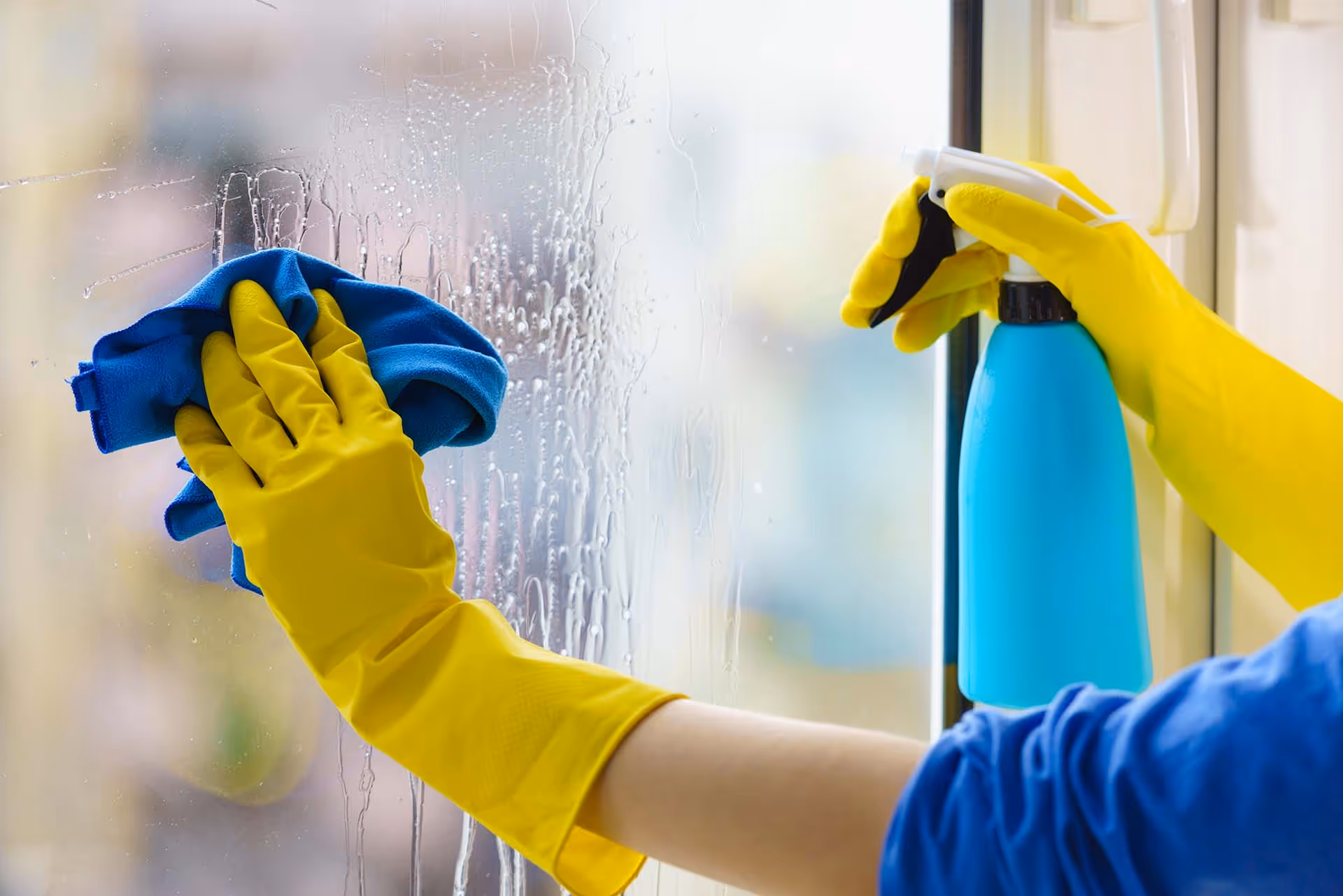 Hemma-avustaja pesemässä ikkunaa keltaisissa kumihanskoissa.