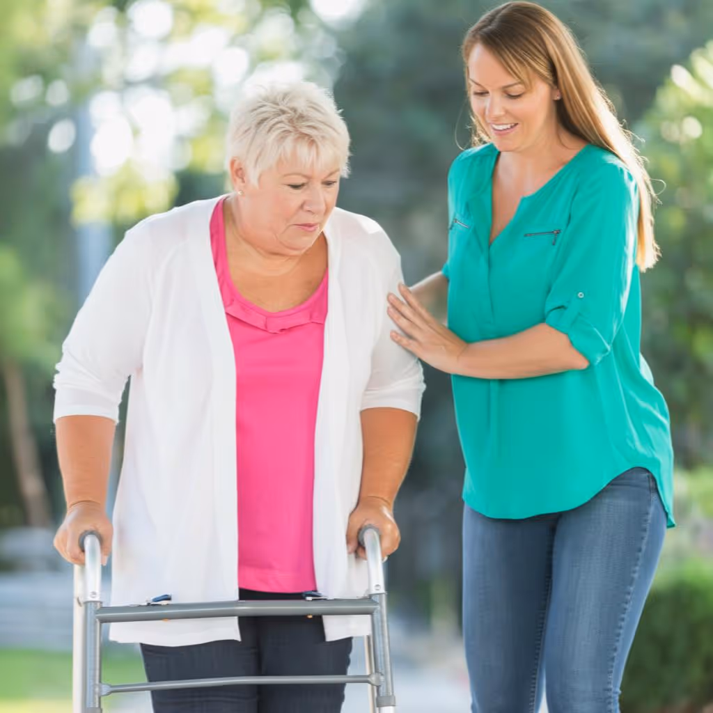 Hemma avustaja tukee ikääntynyttä naista, joka kävelee rollaattorin kanssa ulkona.