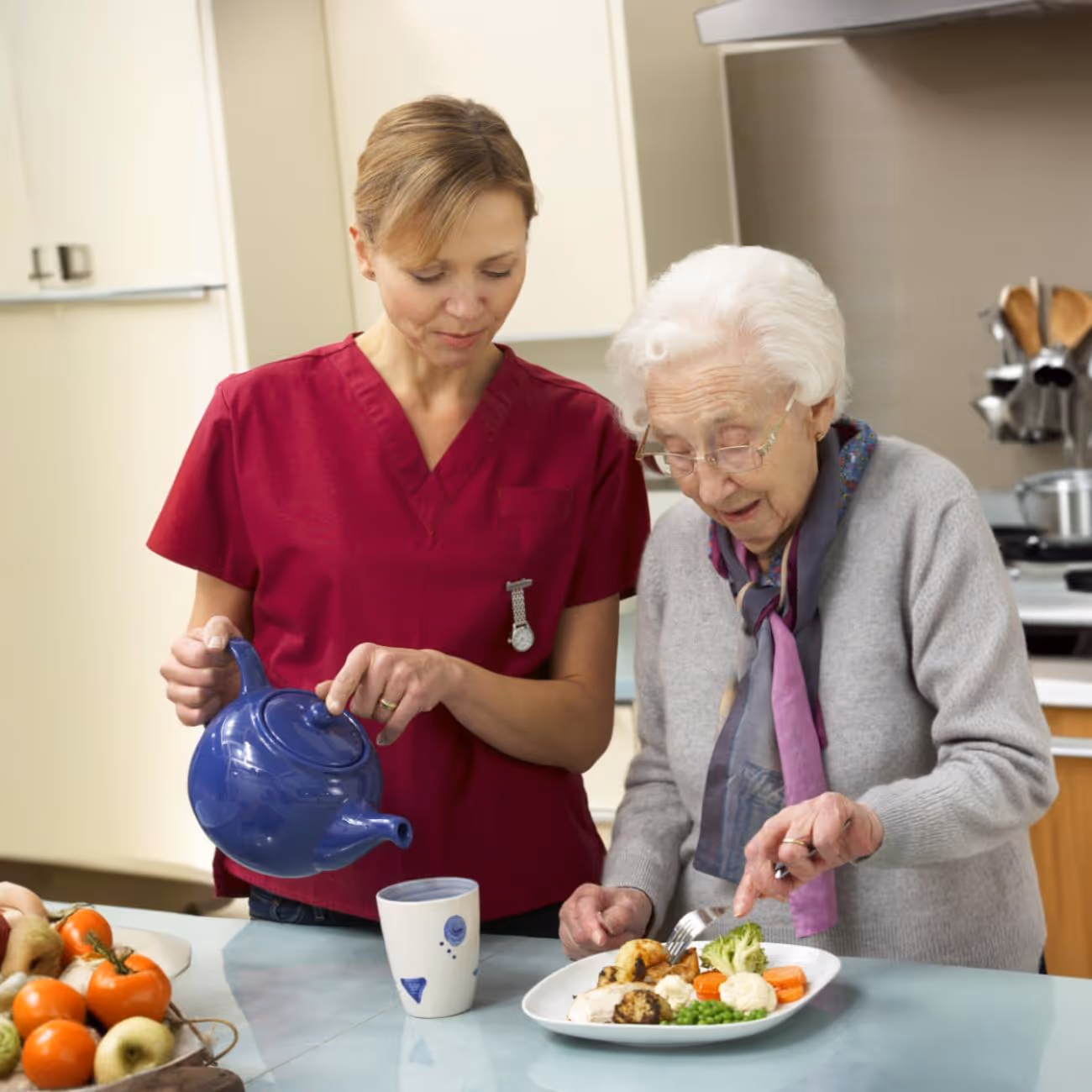 Hemma avustaja kaataa juomaa mukiin iäkkäälle naiselle keittiössä.