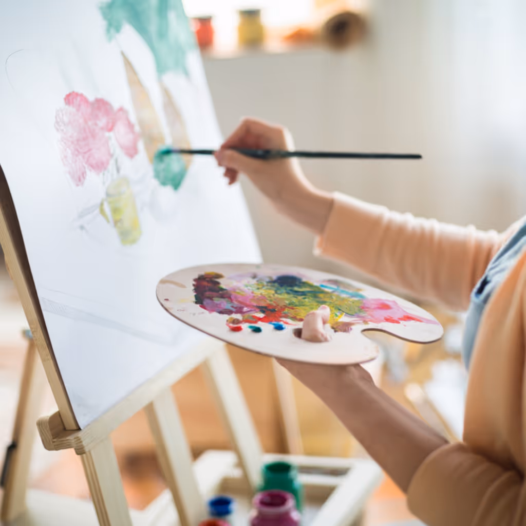 Nainen maalaa kukkia kankaalle käyttäen sivellintä ja maalipalettia.