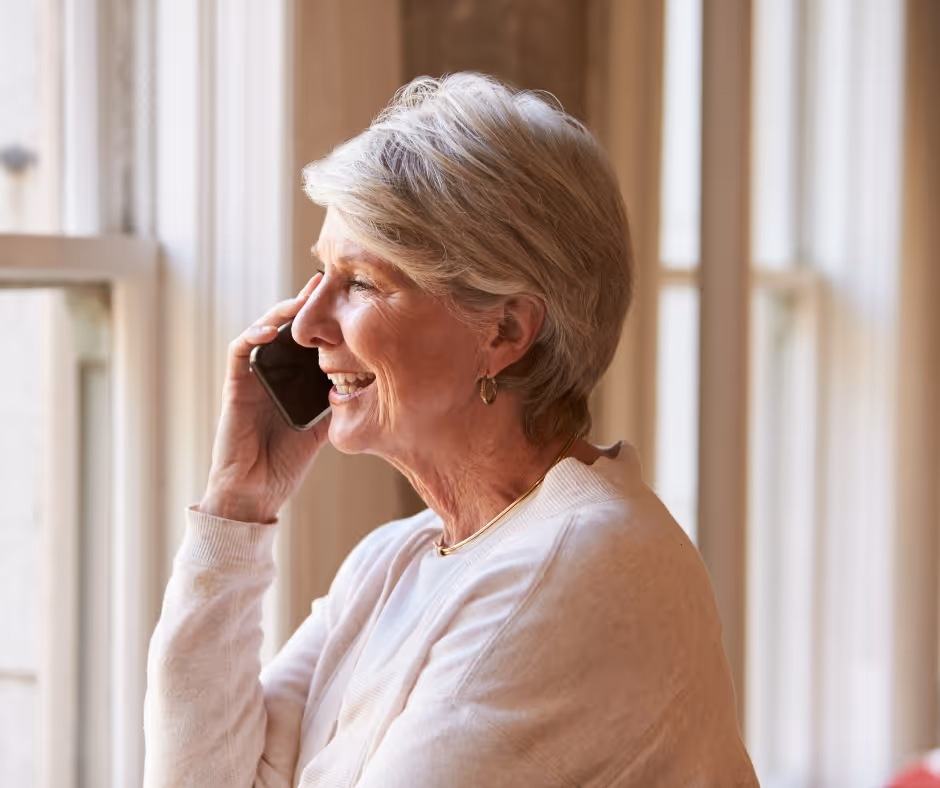 Hymyilevä vanhempi nainen puhuu puhelimessa ikkunan äärellä.