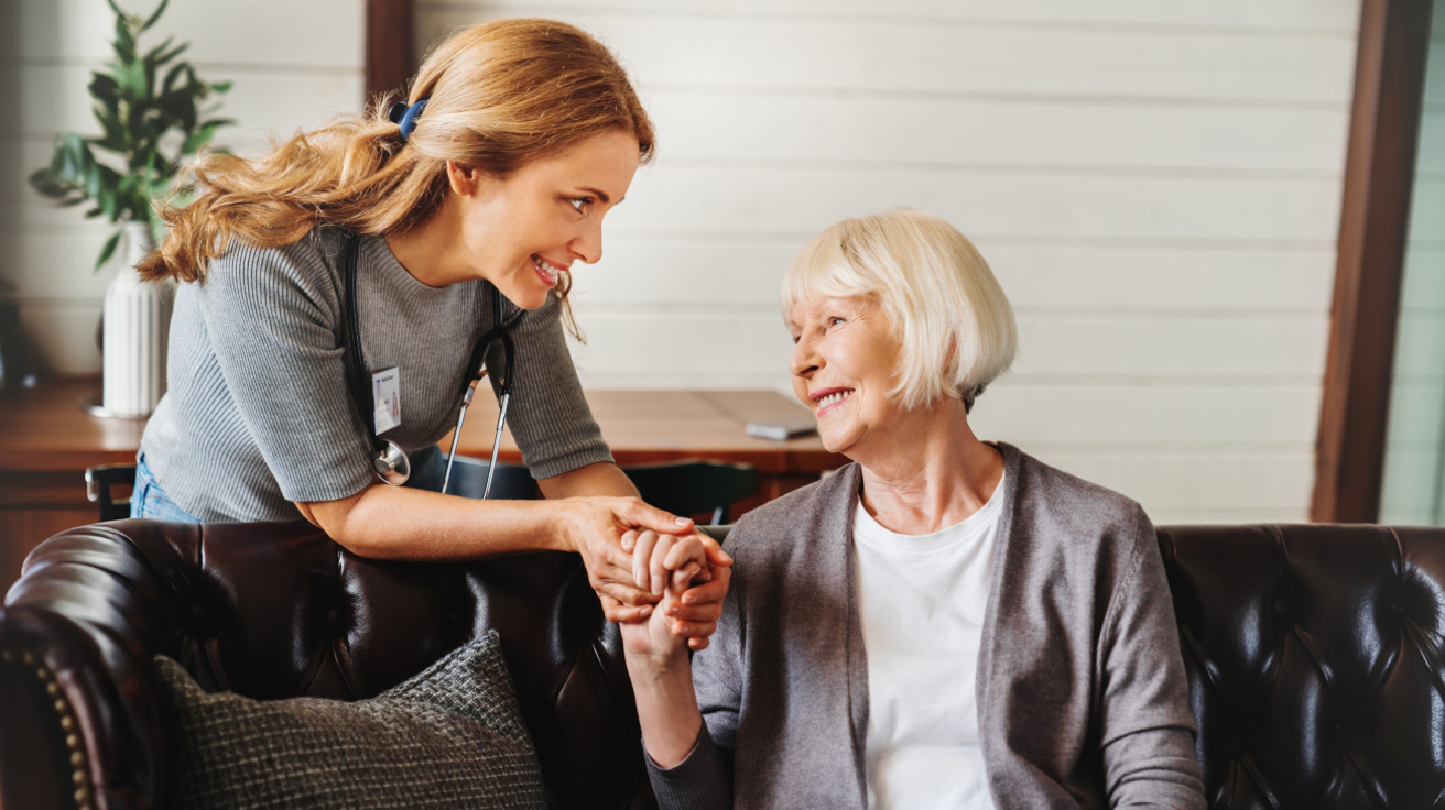 Hemma-avustaja hymyilee ja pitää vanhemman naisen kädestä kiinni.