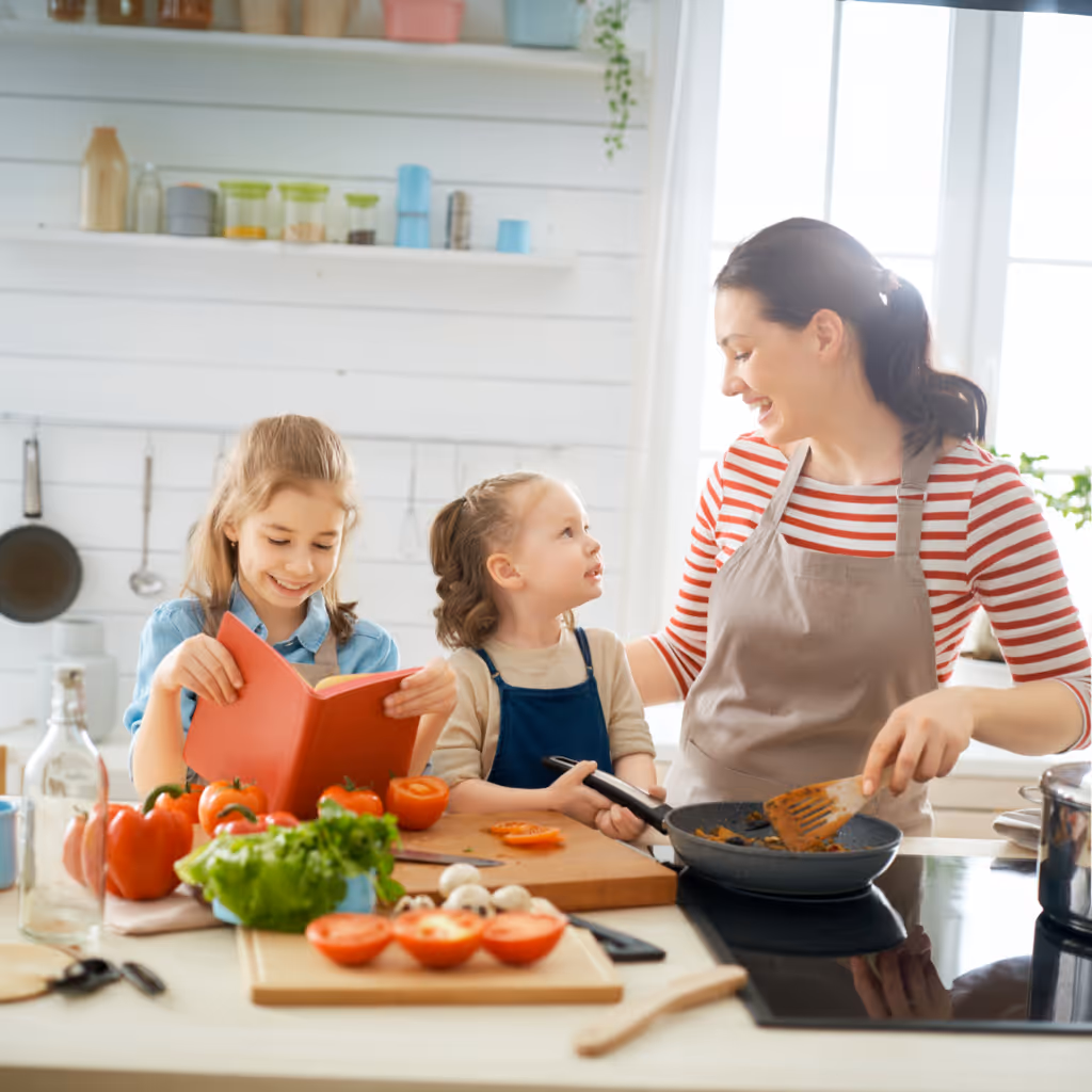 Äiti ja kaksi tytärtä valmistavat ruokaa keittiössä; yksi tyttö lukee reseptikirjaa, toinen katsoo äitiä hymyillen.