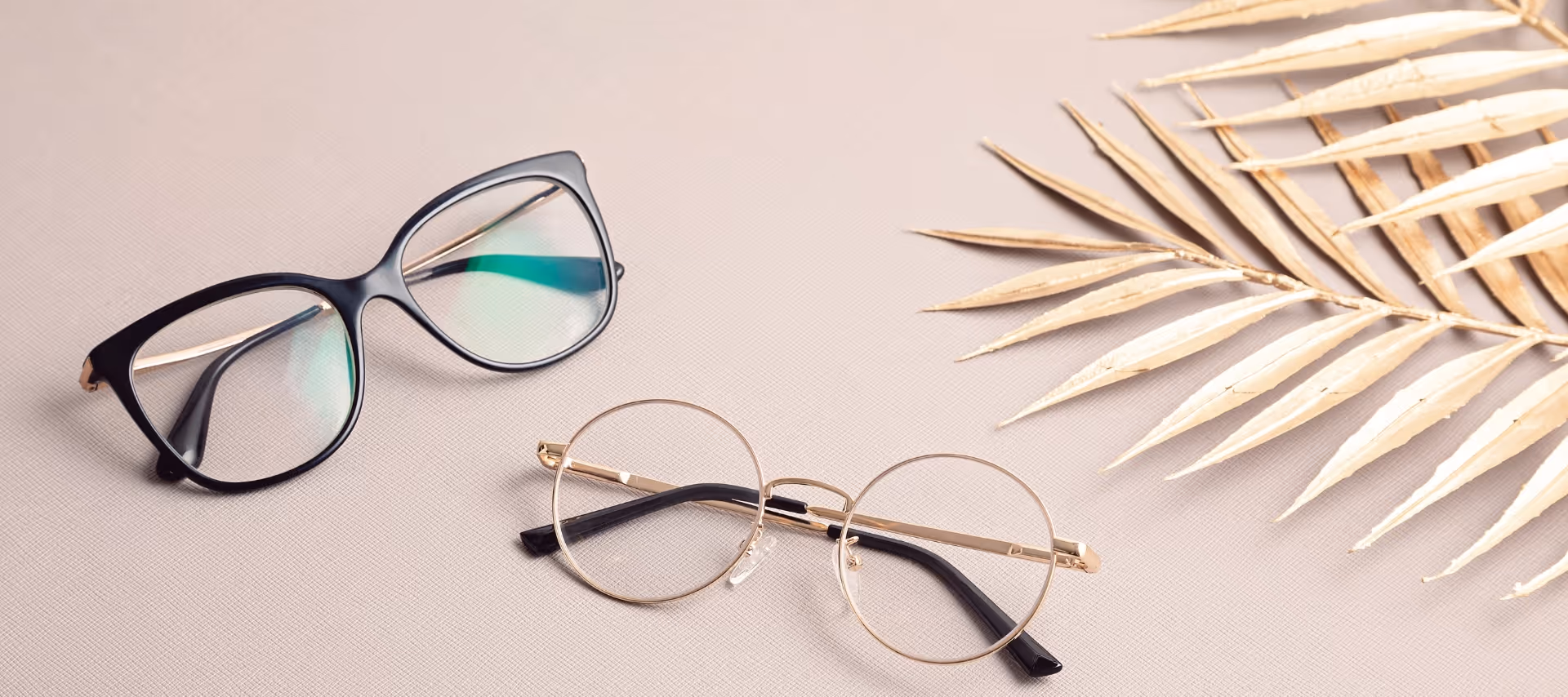 Mustat ja kultaiset silmälasit vaaleanruskealla taustalla, vieressä kuivunut vaalea palmunlehti.