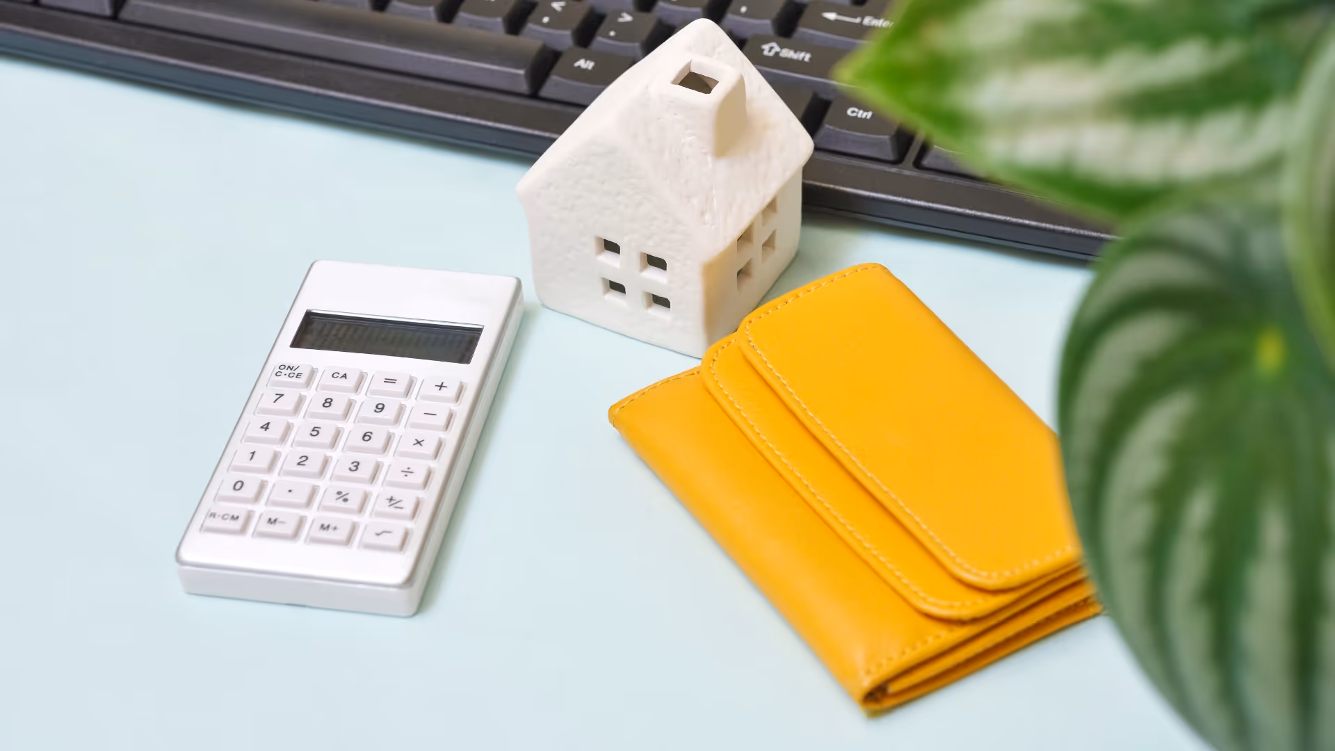 Valkoinen laskin, valkoinen talomalli ja keltainen lompakko vaalealla pinnalla, taustalla osa tietokoneen näppäimistöstä ja vihreitä lehtiä.