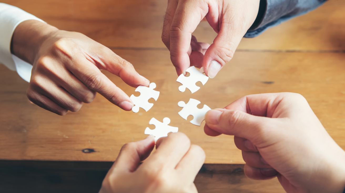 Neljä kättä pitää valkoisia palapelin paloja pöydän päällä.