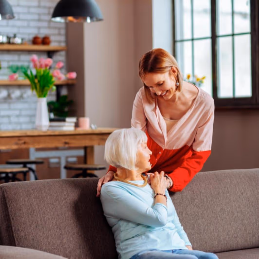 Nuori nainen koskettaa olkapäihin ikääntynyttä naista, joka istuu sohvalla kodikkaassa huoneessa.