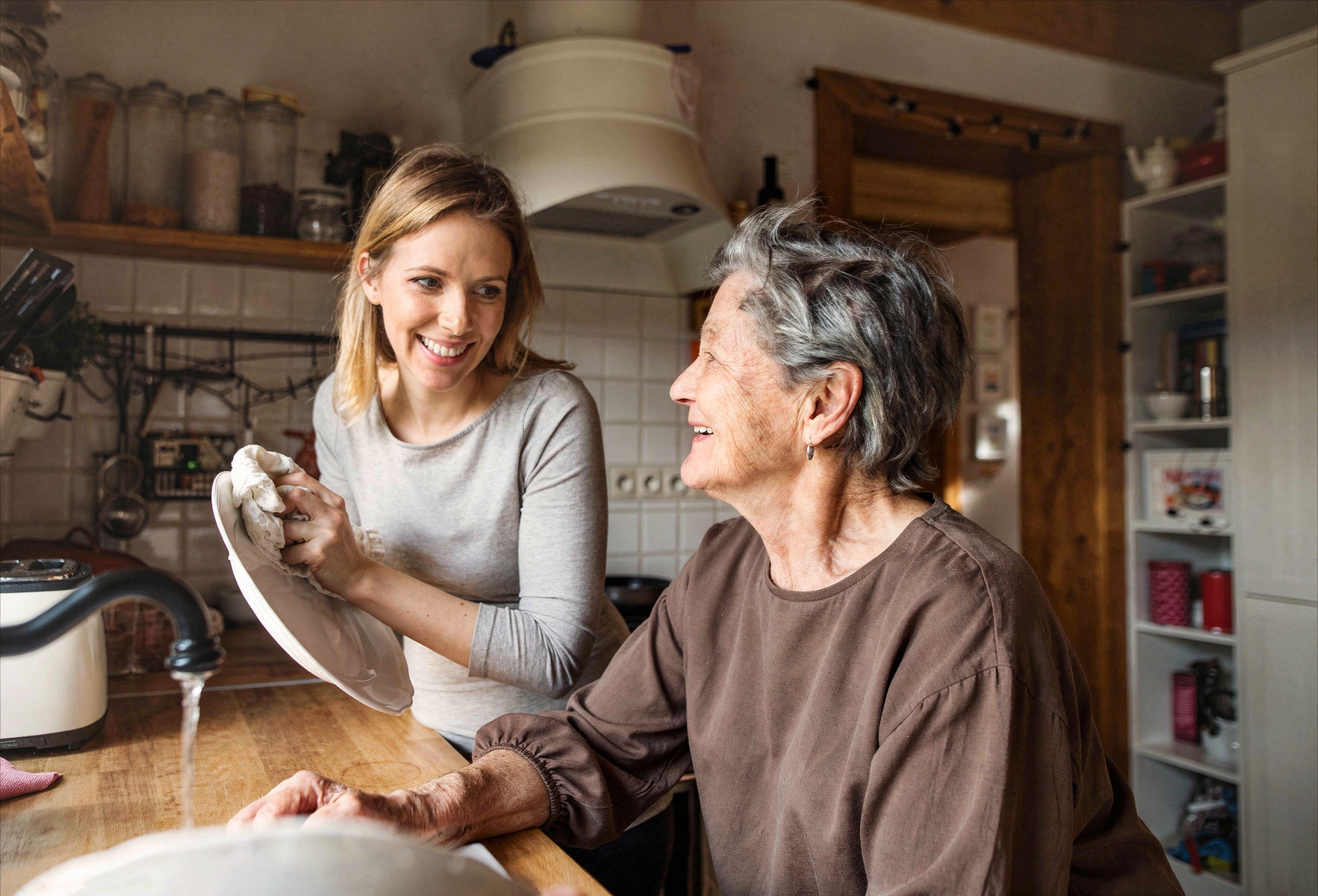 Nuori nainen ja ikääntynyt nainen hymyilevät keittiössä, nuorempi nainen kuivaamassa astioita.