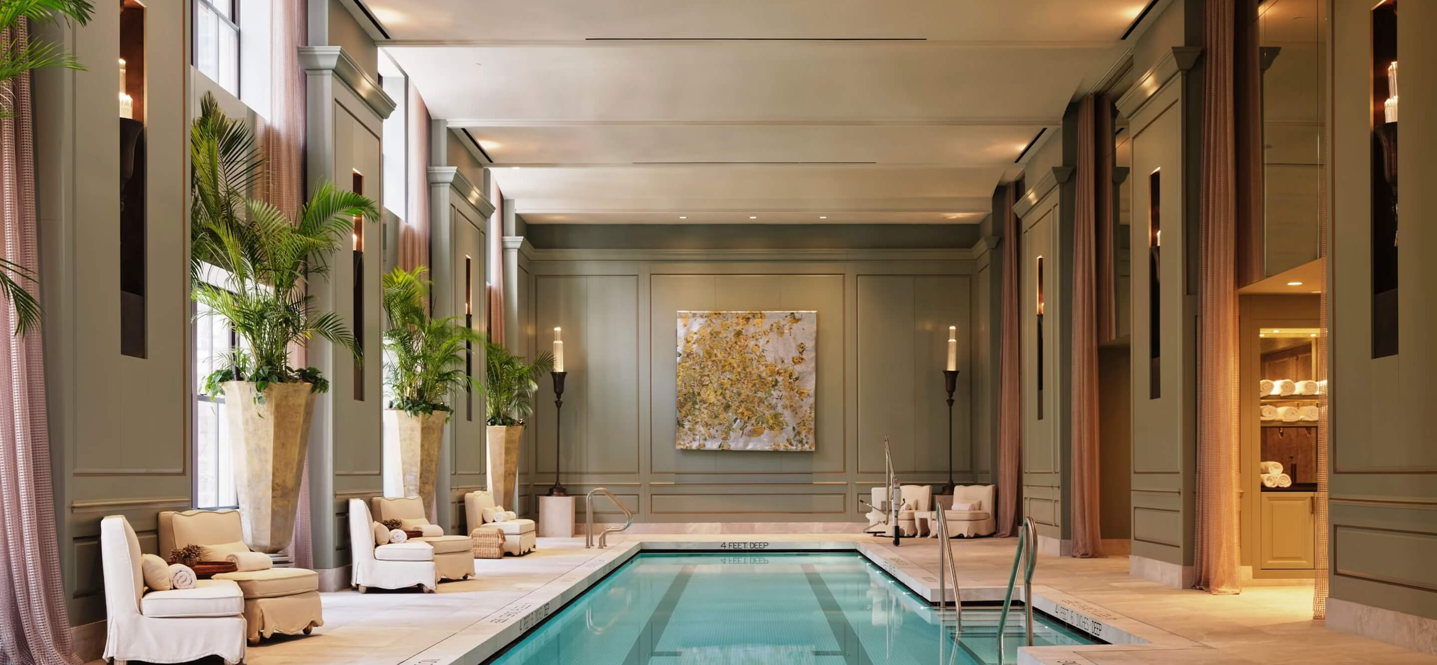 Luxurious indoor swimming pool surrounded by white cushioned chairs, tall potted plants, soft lighting, and elegant wall paneling.