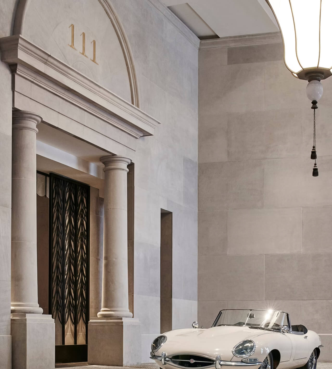 White vintage convertible car parked inside a grand stone building with columns and decorative black metal doors under the number 111.