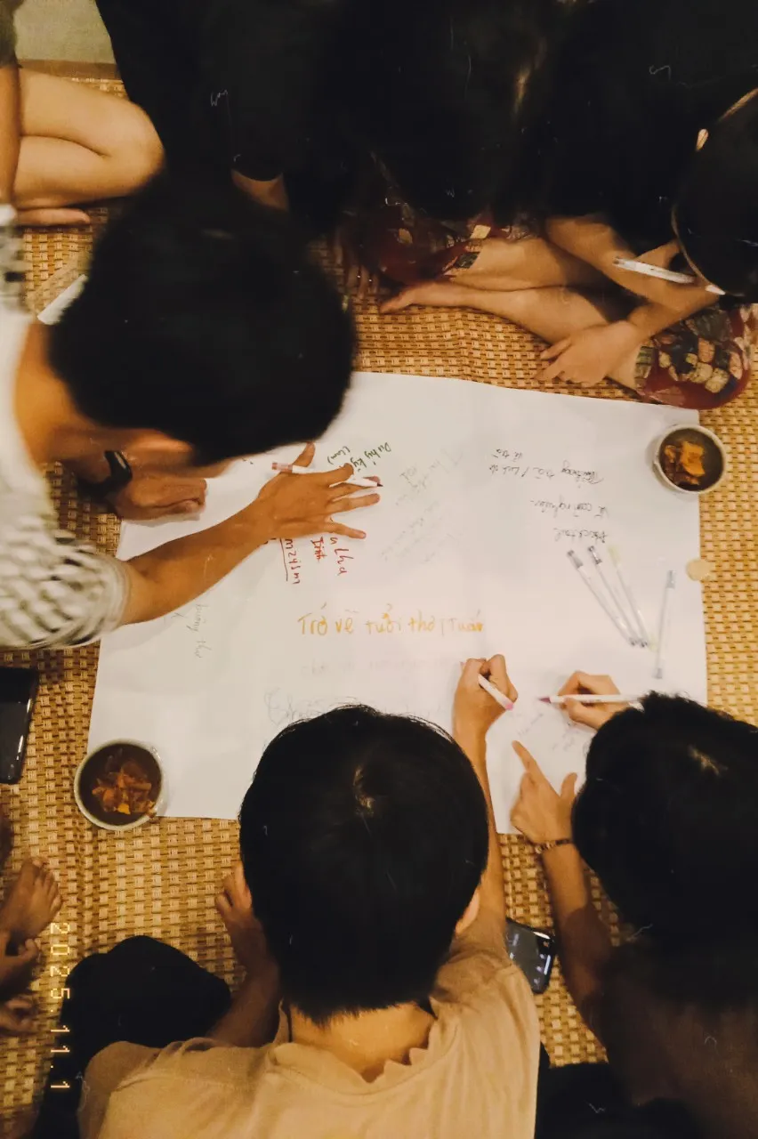 Bốn người phụ nữ đội nón lá và mặc áo phao màu cam đang chèo thuyền trên dòng nước yên bình với cây cối xanh mướt ở ven sông.