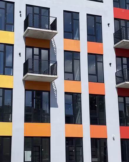 Facade of a modern apartment building with tall windows, small balconies with black railings, and horizontal colored panels in yellow, orange, and red.