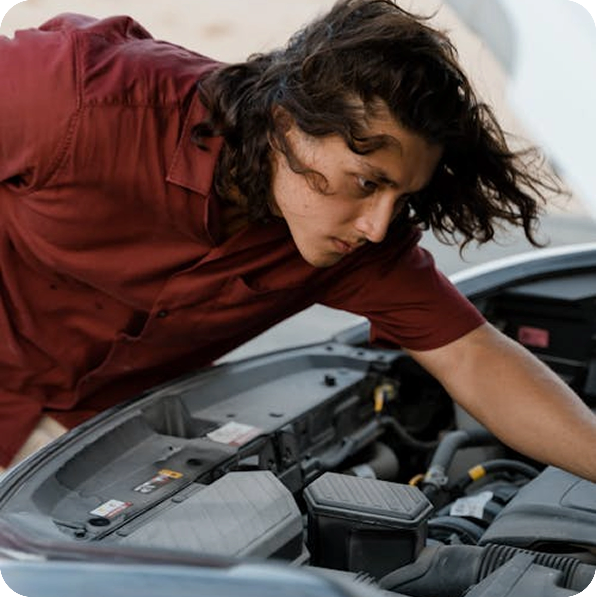 Man with long hair wearing a maroon shirt inspecting the engine of a car with the hood open.