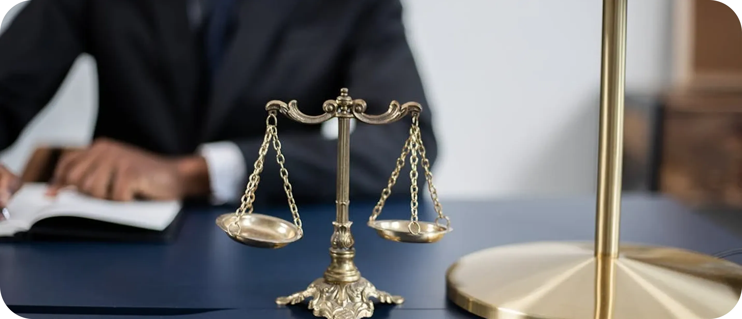 Brass balance scale on a desk with a person in a suit writing in a notebook in the background.