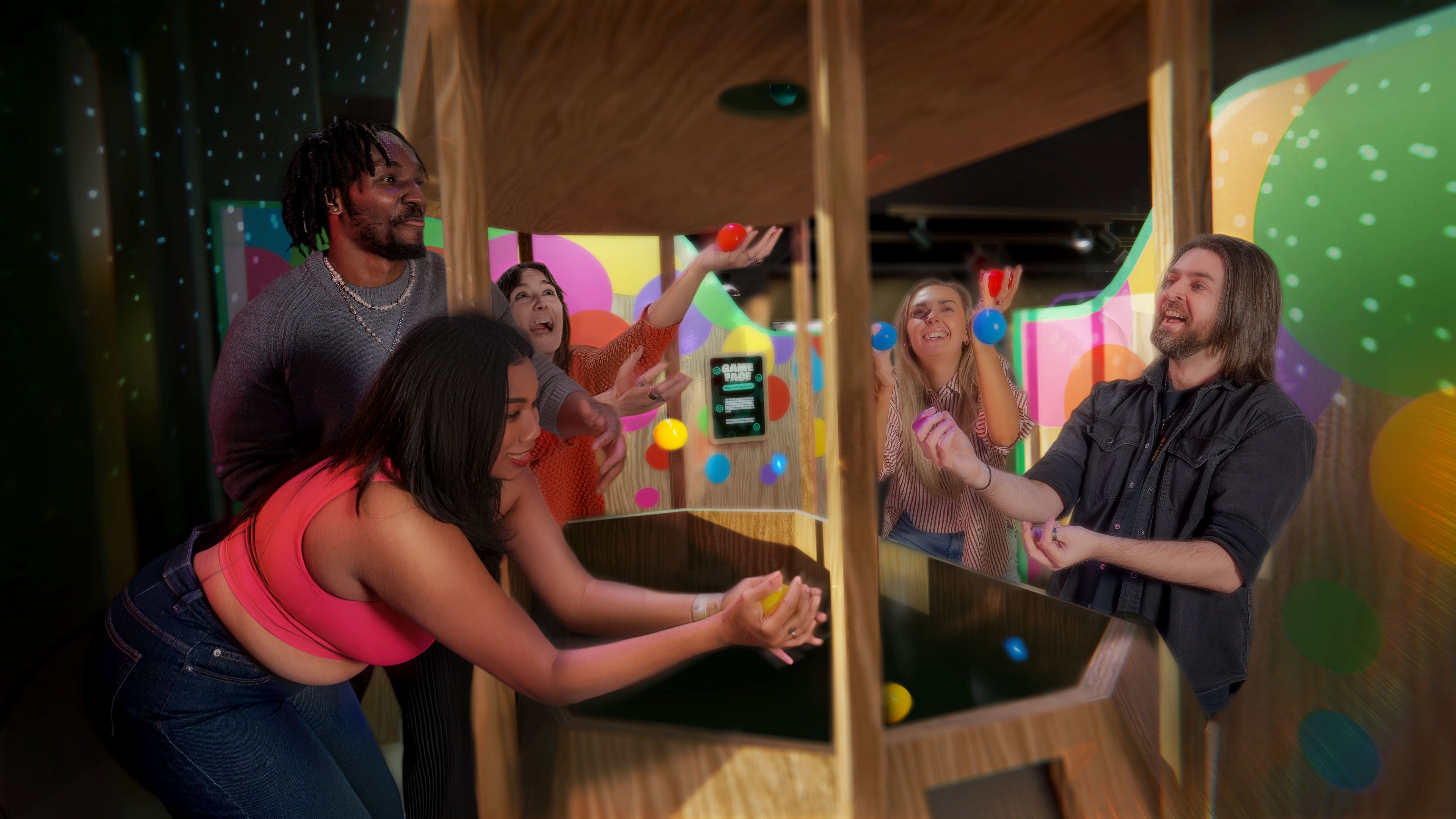 Three people playing in a colorful ball pit; one woman sliding down a yellow slide, another lying in orange overalls, and a man holding a ball to his ear.