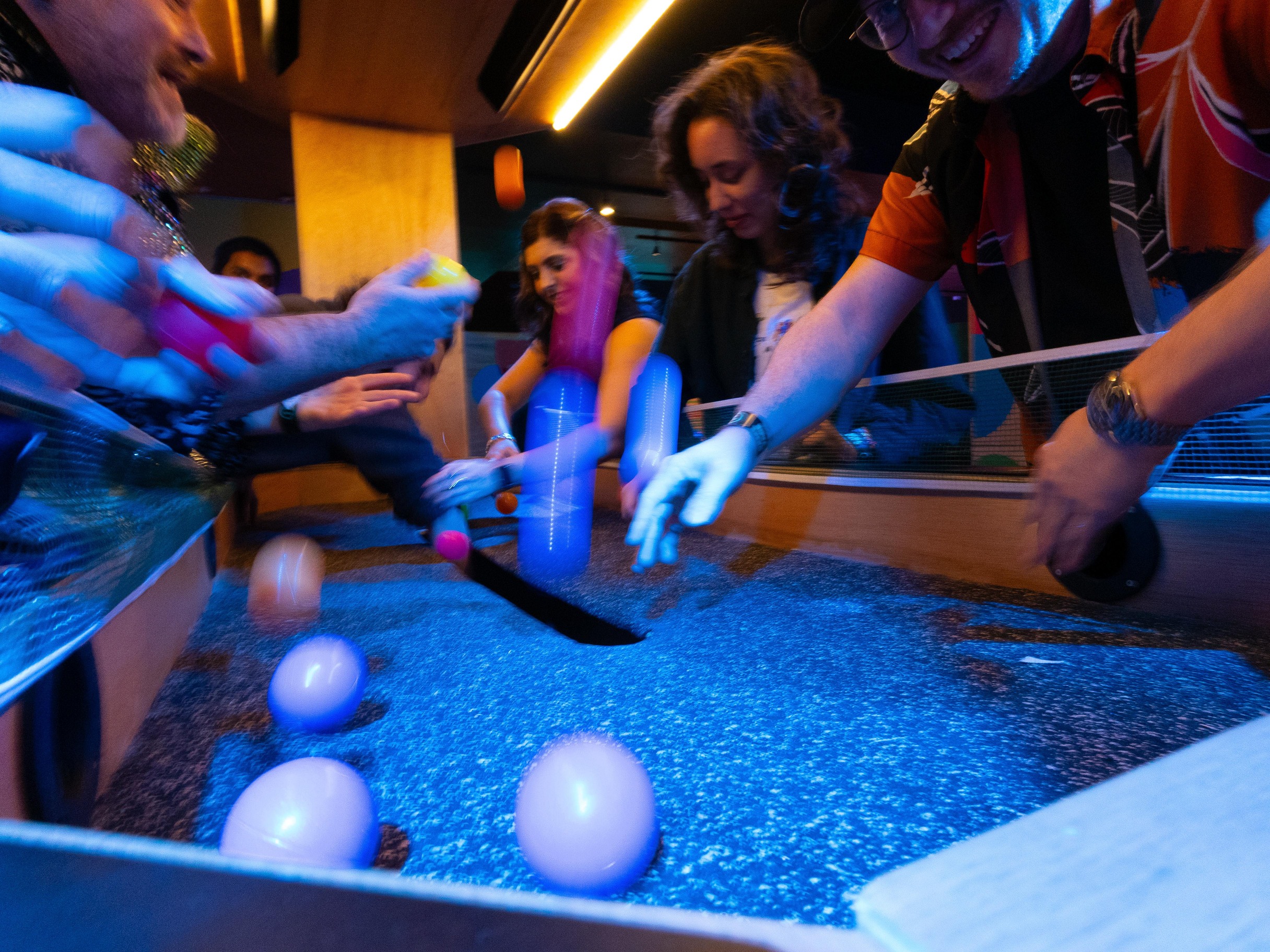 Three people playing in a colorful ball pit; one woman sliding down a yellow slide, another lying in orange overalls, and a man holding a ball to his ear.