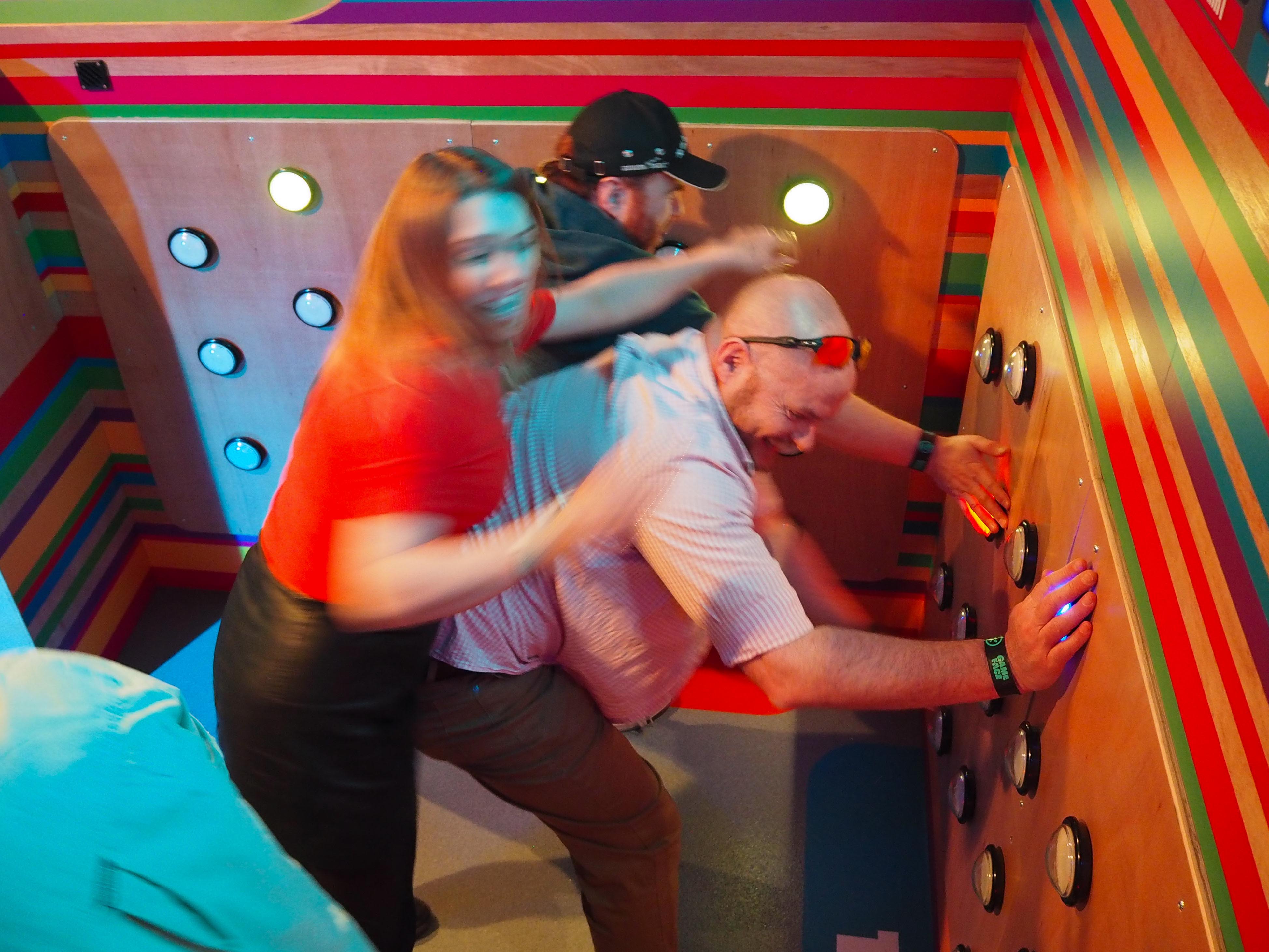 Four young adults laughing and playing with large, round, yellow emoji cushions in a colorful indoor arcade.