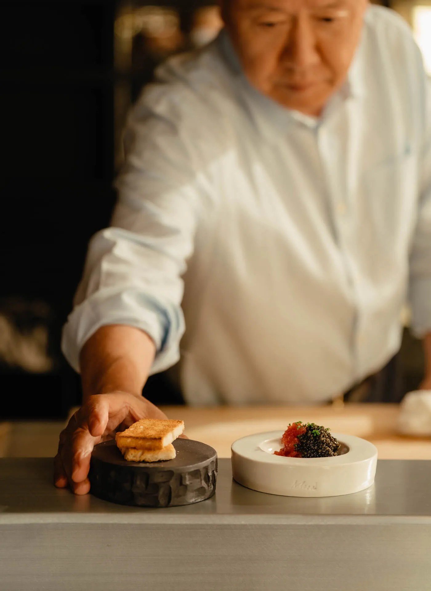 Chef Masa Takayama placing some toasted bread on Masa Design's dish-wear on a counter 
